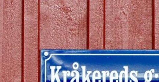 Blåt emaljskylt med Kråkereds g:a skola på faluröd trävägg.