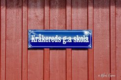 Blåt emaljskylt med Kråkereds g:a skola på faluröd trävägg.